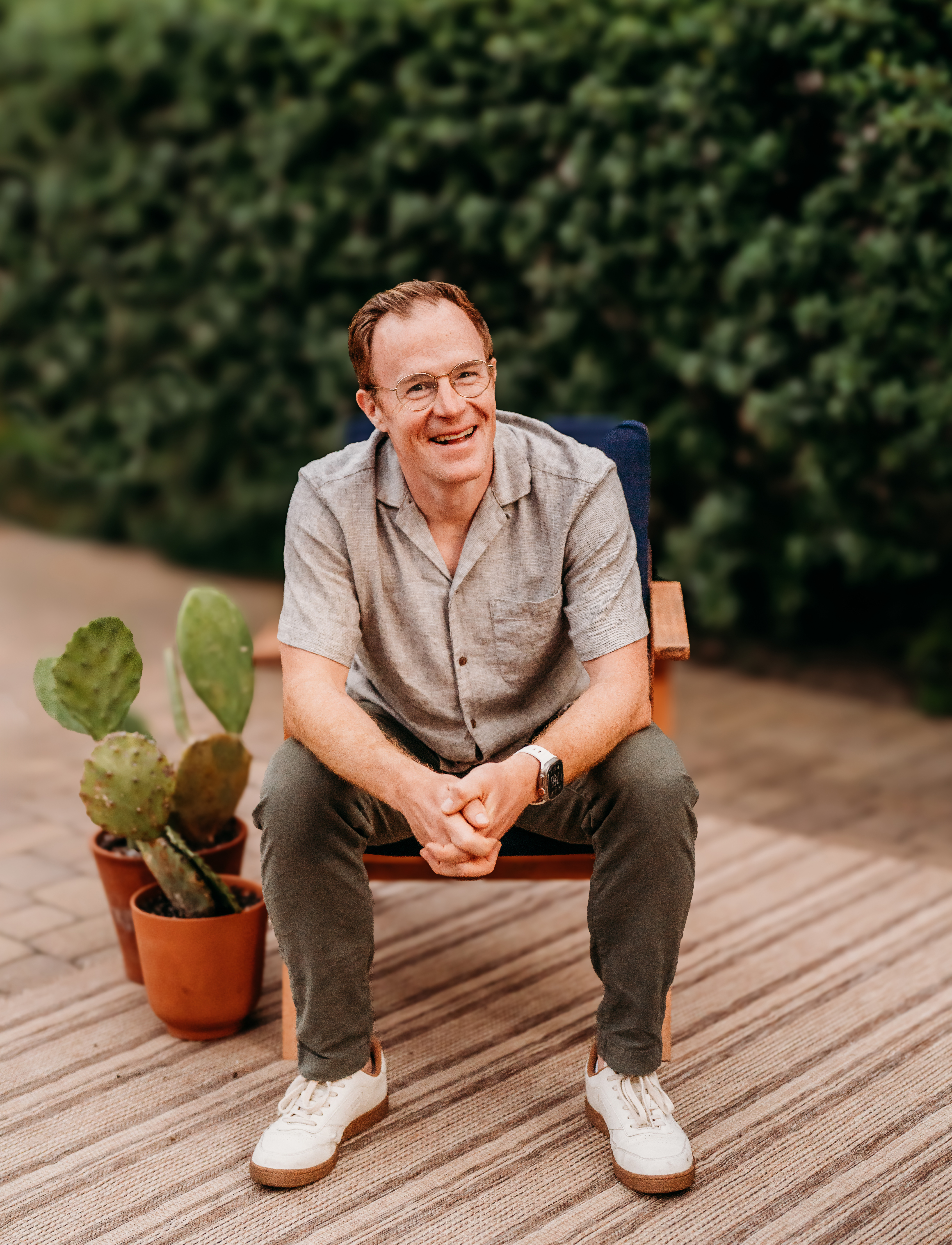 Kyle smiling at the camera, sitting on a chair on a patio with some prickly pear cactus next to him