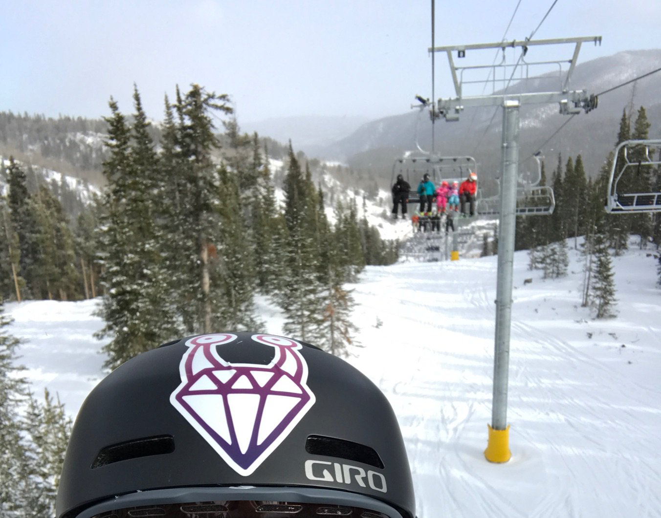 A Keep Ruby Weird sticker on a helmet in a snowy and woody area with a ski lift seat with five people behind