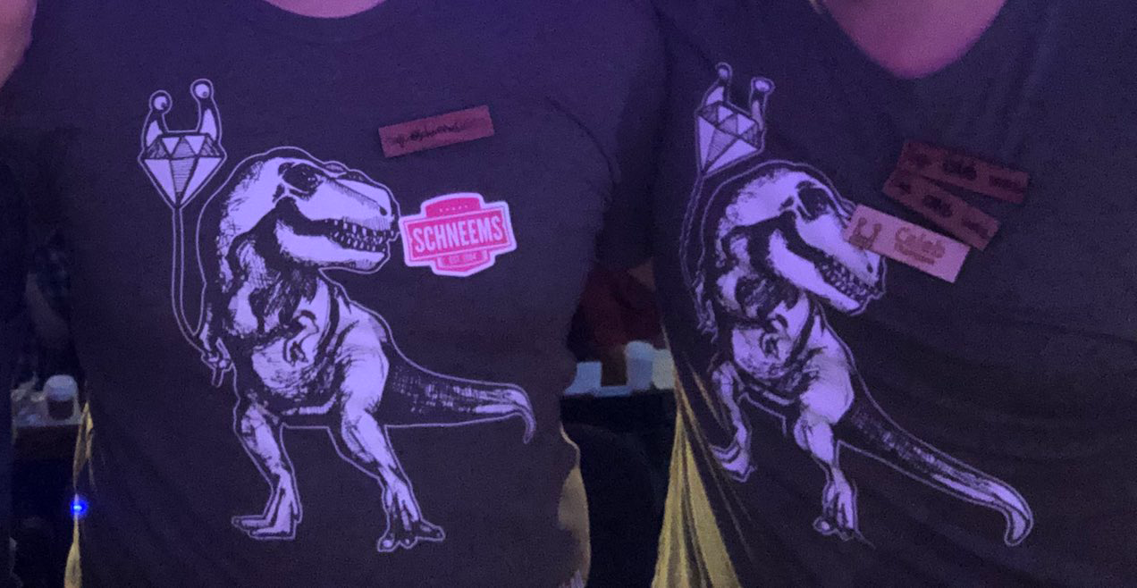 Two people wearing an olive shirt with a graphic of a T-rex holding a Keep Ruby Weird ballon